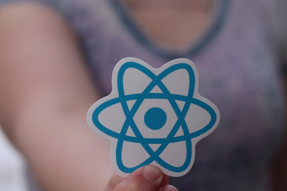 A person holding a sticker featuring a blue atom symbol in front of their chest. The individual is wearing a sleeveless top with a pattern, and the background is blurred, focusing on the sticker. The 