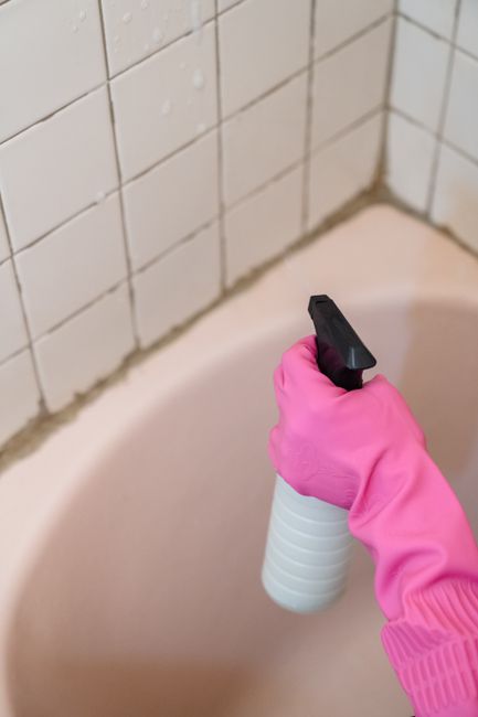 Close-up of a hand wearing a bright pink rubber glove holding a white spray bottle with a black nozzle, positioned above the corner of a bathroom bathtub. The background features white ceramic tiles o